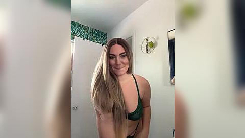 Media: Video of a smiling young woman with long blonde hair, wearing a green bra, standing in a bathroom with white walls and a green patterned shower curtain.