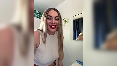 Media: Video of a smiling, light-skinned, blonde woman with red lipstick, wearing a white, sleeveless top, in a small, white-walled room with a mirror and a blue curtain.