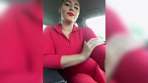 Media: Video of a confident woman with fair skin and red lipstick, wearing a tight, bright red tracksuit, sitting in a car, smiling.