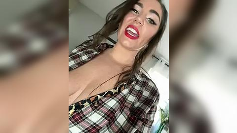 Media: Video of a smiling, light-skinned woman with long brown hair, wearing a low-cut plaid shirt, her cleavage visible, indoors with blurred background.