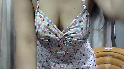 Media: A close-up video of a woman's torso in a colorful, polka-dotted sundress with thin straps, revealing ample cleavage. Background shows beige curtains and a wooden chair.