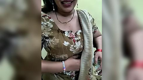 Media: Video of a smiling South Asian woman wearing a floral saree, with a beige shawl draped over her shoulders. She has a red bindi and multiple bangles on her wrist. The background is blurred, focusing on her joyful expression.