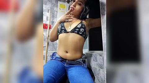 Media: Video of a South Asian woman with medium skin tone and medium-sized breasts, wearing a black bikini top with white skulls and blue jeans, sitting in a gray-tiled bathroom with medical equipment in the background.