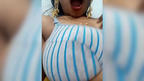 Media: Video of a woman with dark skin, wearing a blue and white striped tank top, large gold earrings, and a gold necklace, mouth open in excitement.