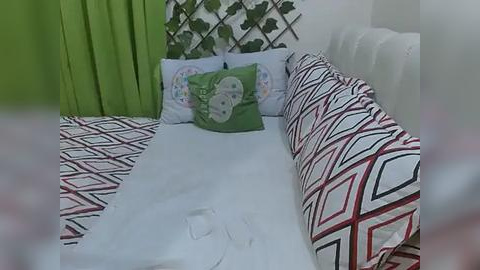 Media: A video of a small, cozy bedroom with a white bed adorned with a geometric-patterned duvet cover and pillows. The background features a green curtain, a white wall with a plant, and a leafy vine pattern.