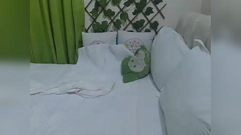 Media: Video of a cozy bedroom with white bedding, green curtains, and a leaf-patterned wall. The bed is made with a floral pillow, and there's a discarded white garment on the floor.