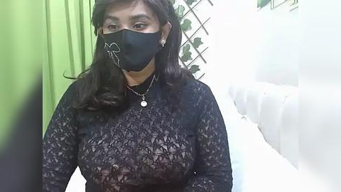 Media: A video of a woman with medium brown skin, long dark hair, wearing a black lace top, black face mask, and a necklace, standing in a brightly lit room with green and white walls.