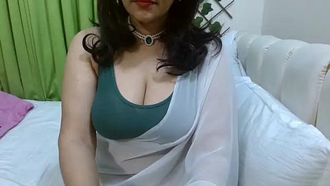 Media: Video of a light-skinned woman with long, wavy black hair, wearing a green tank top and a white draped shawl, sitting on a white leather couch in a room with green and purple curtains.