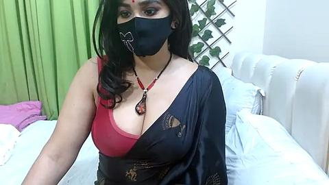 Media: Video of a South Asian woman with medium skin tone, wearing a black mask, black saree, red bra, and red necklace, sitting on a white bed in a cozy room with green curtains.