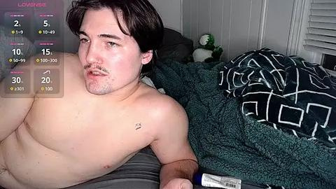 Media: Video of a shirtless, slightly overweight, light-skinned man with dark hair lying on a bed, wearing a blue wristband, next to a geometric-patterned blanket.