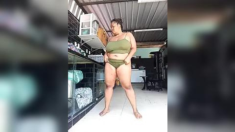 Media: Video of a plus-size Latina woman in olive green bikini, standing in a dimly lit, cluttered storage room with shelves and a white AC unit.