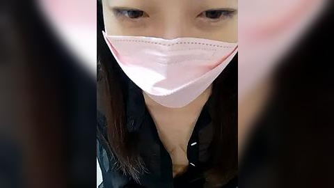 Media: Video of a close-up of an Asian woman wearing a pink surgical mask, with long dark hair, in a blurred, dimly-lit room.