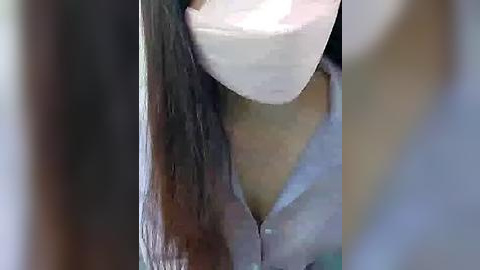 Media: Video of a person with long brown hair, wearing a white surgical mask, and a light blue top, seen from a low angle, creating a blurred, abstract background.