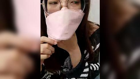 Media: Video of a woman with long brown hair, wearing glasses, a black and white patterned sweater, and a pink surgical mask, slightly blurry in the background.