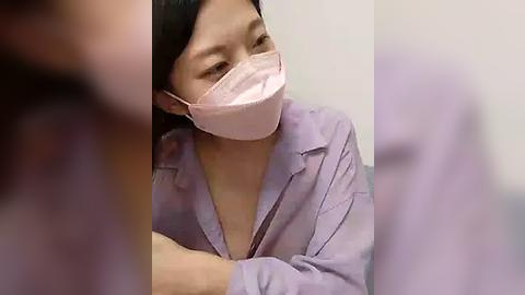 Media: Video of an Asian woman with straight black hair, wearing a light pink surgical mask and a lavender hospital gown, smiling, seated in a hospital setting.