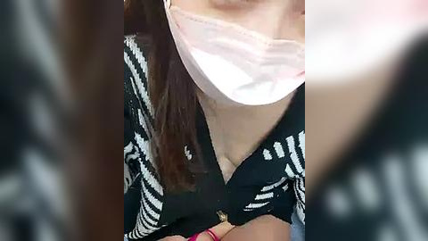 Media: Video of a person with long dark hair wearing a white surgical mask, black and white striped shirt, and pink fingernails, partially blurred background.