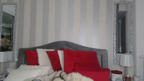 Media: Video of a chic, modern bedroom featuring a gray tufted headboard, red and white pillows, and a white fur blanket. The background has striped white and gray wallpaper, and large silver-framed mirrors flank the bed.