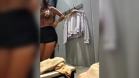 Media: Video of a woman in a dressing room, wearing a sheer, brown top and black shorts, holding a white shirt, with beige knitted sweaters on a bench.