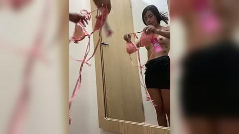 Media: Video of a young woman with medium skin tone, straight black hair, in a black skirt, adjusting a pink bra in front of a mirror. Pink streamers are visible in the background.