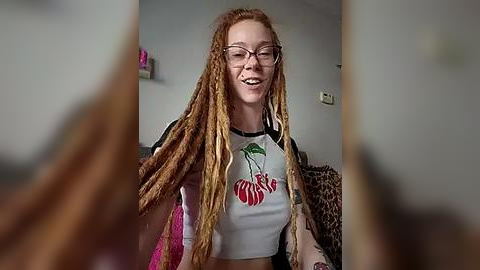 Media: Video of a smiling woman with long, dreadlocked hair, wearing glasses, a white crop top with cherry graphic, and a leopard print jacket. Background shows a beige wall and a pink object.