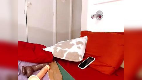 Media: A video of a small, cozy bedroom with red walls and a red bed, featuring a white pillow with cow spots, a smartphone, and a wooden dildo on a green bedspread.