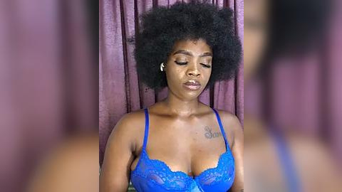 Media: Video of a black woman with a large, natural afro, wearing a blue lace bra, standing in front of mauve curtains. Her eyes are closed, and she appears serene.