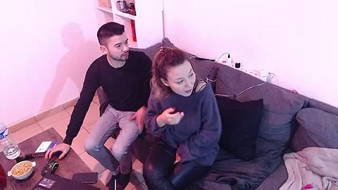 Media: Video of a young couple, a man with short dark hair in a black sweater and jeans, and a woman with brown hair in a dark sweater, watching TV on a gray couch.