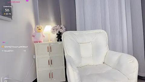 Media: A video of a cozy, minimalistic bedroom with a white armchair, a small white dresser, a fluffy pink duck toy, and a vase of pink flowers beside a lit lamp.