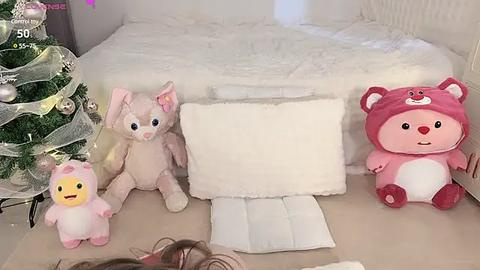 Media: Video of a cozy bedroom scene with a plush pink bear, white pillow, and pink bunny plushies near a white bed. Christmas decorations and a soft blanket in the background.