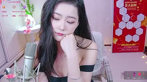 Media: A video of an Asian woman with long black hair, wearing a black off-shoulder top, leaning on a microphone, in a pink-themed room with posters.