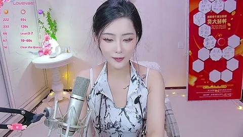 Media: A video of an Asian woman with fair skin, dark hair in a bun, wearing a white halter top, sitting at a desk with a microphone, flowers, and a calendar on a wall in the background.