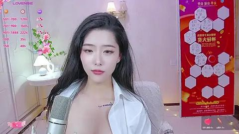 Media: A video of a young East Asian woman with long black hair, wearing a white button-down shirt and a gray blazer, posing indoors with a pink flower vase and a red banner in the background.