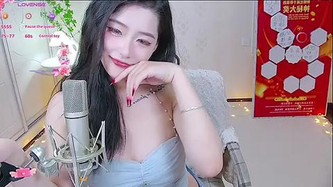 Media: Video of an Asian woman with long black hair, light skin, and red lipstick, wearing a light blue off-shoulder top, sitting at a desk with a microphone, smiling. Background includes a red poster and greenery.