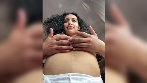 Media: Video of a pregnant woman with curly hair, wearing a white top, hands on her large belly, blurry background.