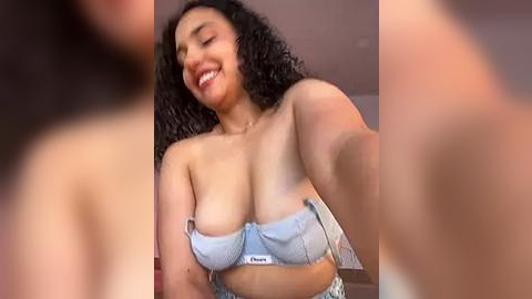 Media: A video of a smiling, medium-skinned woman with curly hair, wearing a light blue bra and jeans, taken from a slightly blurred, close-up angle.