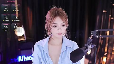 Media: Video of a young, fair-skinned East Asian woman with light brown hair in a loose bun, wearing glasses and a light blue shirt, speaking into a microphone against a dark, studio-lit background.