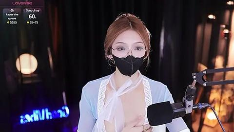 Media: Video of a woman with glasses, wearing a black face mask and light blue blouse, speaking into a microphone on a stage with dimly lit, blurred background.