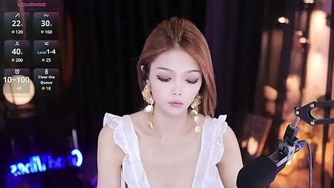 Media: Video of an Asian woman with long, straight brown hair wearing a white ruffled dress, recording on a black microphone. She has fair skin, wearing gold dangling earrings. Background is blurred with studio lights.