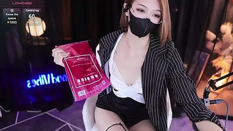 Media: Video of an Asian woman with light skin, wearing a black face mask, pinstriped blazer, white lace bra, and black shorts, holding a pink \"SANTA\" gift bag, sitting on a white chair in a dimly lit room with a blue LED strip.