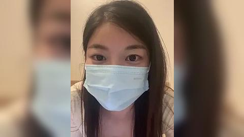 Media: Video of an East Asian woman with long black hair, wearing a light blue medical mask, looking directly at the camera. She is in a light-colored shirt. The background is blurred.