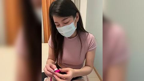 Media: A video of an Asian woman with long black hair, wearing a pink short-sleeved shirt, white mask, and headphones, meticulously applying nail polish on her fingers in a dimly lit room with a wooden door and beige walls.