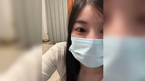 Media: Video of a young Asian woman with long black hair, wearing a light blue face mask, and a beige top. She is indoors with a white curtain in the background.