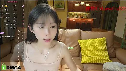 Media: A video of an East Asian woman with long black hair, wearing a beige spaghetti-strap top, sitting on a brown couch in a cozy living room.