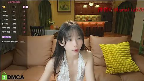 Media: A young Asian woman with long black hair, wearing a white lace top, sits on a brown sofa in a modern living room with a kitchen backdrop.