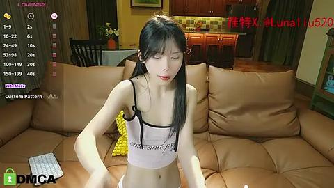 Media: A video of an East Asian woman with long black hair, wearing a white tank top, sitting on a brown couch in a cozy living room.