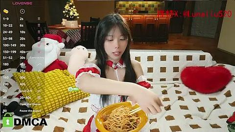 Media: A video of an Asian woman with long black hair, wearing a red and white Santa outfit, eating noodles on a patterned couch. The background shows a modern kitchen.