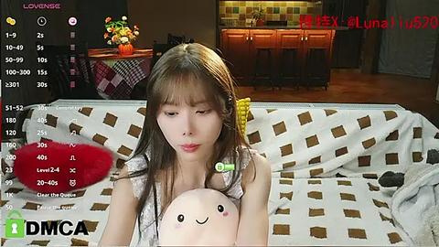 Media: Video of a young Asian woman with straight brown hair, wearing a white floral dress, cuddling a stuffed toy bear on a patterned couch. Background shows a cozy living room with a kitchen area.