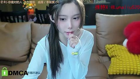 Media: A video of an Asian woman with long black hair, wearing a white blouse, sitting on a beige couch, biting her finger in a casual indoor setting.