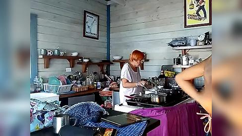 Media: A video of a rustic kitchen with wooden walls, featuring a red-haired woman in a white shirt preparing food on a purple counter. Shelves hold dishes and a poster reads \"Coca-Cola.\