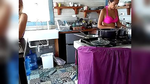 Media: Video of a woman in a vibrant pink bikini cooking on a stove in a rustic kitchen with mismatched decor, including a blue water jug and a white sink.